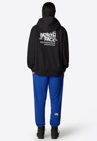 Zwarte hoodie met wit logo op de achterkant, gecombineerd met blauwe joggingbroek. Geweven stof, comfortabele pasvorm en zwarte schoenen met grijze accenten.
