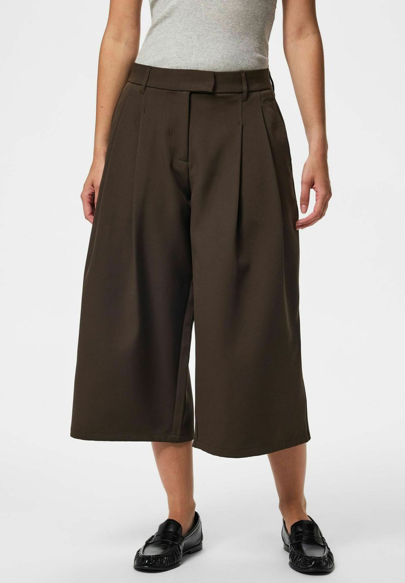 Femme portant un pantalon court large marron foncé, un haut gris sans manches et des mocassins noirs, debout devant un fond blanc.