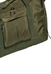 Olijfgroene duffelbag van duurzaam materiaal, met een voorzak met ritssluiting, stevige handvatten en een bronzen logo-accent.