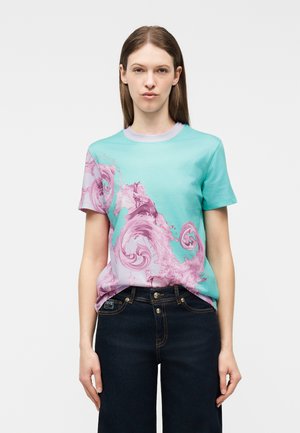 Femeie cu păr drept, maro, purtând un tricou verde mentă cu modele roz în formă de spirale și blugi albastru închis, pe un fundal simplu.