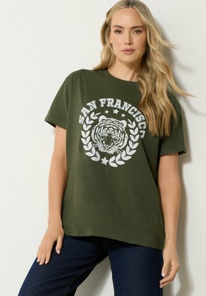 Blondine draagt een donkergroen t-shirt met San Francisco-opdruk, tijgerkop en lauwerkrans, gecombineerd met donkerblauwe spijkerbroek, tegen een lichte muur.