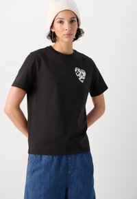 T-shirt nera a maniche corte in cotone con una grafica a forma di cuore bianca sul petto. Indossata con pantaloni di jeans blu a taglio largo.