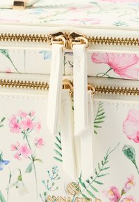 Valkoinen käsilaukku, jossa kukkakuvio ja kultaiset vetoketjut sekä teksturoitu pinta. Koristeena vaaleanpunaisia, sinisiä ja vihreitä kukkia sekä brändin logo.