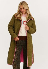 Dlhý prešívaný kabát olivovo zelenej farby s ružovým lemovaním, vybavený predným zipsom a cvokmi, nosený cez vzorovaný sveter a nohavice so zvieracím motívom.