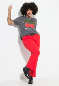 T-shirt graphique en gris charbon avec un motif de cerises rouges, associé à un pantalon évasé rouge vif et des bottines noires à clous décoratifs.