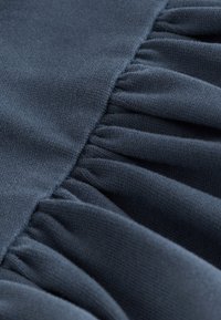 Tmavě modrá tkanina s jemnou texturou, která na jedné straně zdobí nashromážděné volánky, vyzdvihující jemný a hladký design.