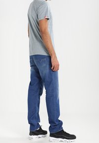 Harmaa lyhythihainen t-paita, siniset farkut, mustat ja valkoiset lenkkarit. Ominaisuuksiin kuuluu logo hihassa, rento istuvuus ja suora lahje.