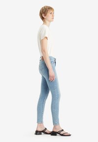 Világoskék, slim-fit farmernadrág, fehér rövidujjú inggel és fekete magassarkú szandállal párosítva. Oldalnézet, minimális részlet.