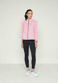 Veste zippée rose clair avec un motif texturé, associée à un legging noir et des baskets blanches avec des accents bleus.