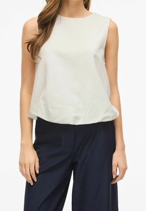 Femme portant un chemisier blanc sans manches et un pantalon bleu foncé, debout devant un fond uni.