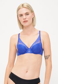 Blå blonde-bh med justerbare stropper, med et dekorativt design og bølgete kanter. Inkluderer en liten metallisk detalj i midten.
