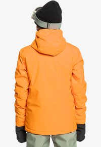 Veste imperméable orange vif avec une capuche, dotée de manches longues. La personne porte des gants noirs. L'arrière-plan est blanc uni.