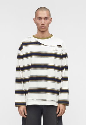 Άνδρας με ξυρισμένο κεφάλι που φορά υπερμεγέθη λευκή μπλούζα με οριζόντιες ρίγες σε ναυτικό μπλε και ελιά και μαύρο παντελόνι, στέκεται μπροστά σε απλό φόντο.