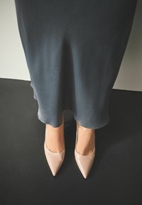 Beežid lakknahast terava ninaga kontsakingad, mida on kombineeritud voolava tumeda halli midi kleidiga, taustal tume põrand ja hele sein.