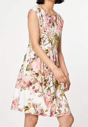 Vrouw draagt een mouwloze, knielange witte jurk met roze en groene bloemenprint, staand tegen een effen witte achtergrond.