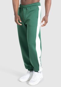 Mand iført grønne joggingbukser med hvide sidelinjer og hvide sneakers, stående foran en ensfarvet baggrund.