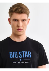 Mladić sa kratkom svijetlosmeđom kosom, odjeven u crnu majicu s plavim natpisom "BIG STAR" i bijelim natpisom "Your Life. Your Jeans." na bijeloj pozadini.