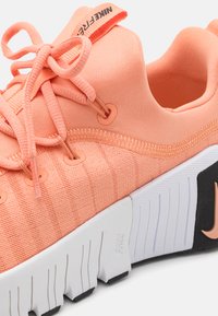 Korallni Nike Free teniske z minimalnim teksturiranim materialom, ki imajo ukrivljeno vezanje, izrazite oranžne detajle in črno-bele podplate.