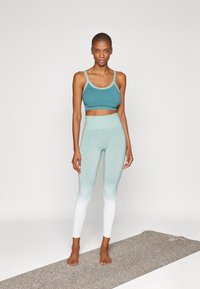 Sujeción deportiva color verde azulado con tirantes delgados, combinada con leggings ombre en degradado que van de verde azulado a blanco. La modelo está descalza sobre una colchoneta de yoga.