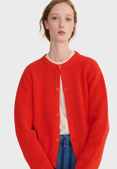 Jeune personne à la peau claire et aux cheveux roux portant un cardigan en maille épaisse orange vif à boutons, par-dessus un haut blanc et un jean bleu.