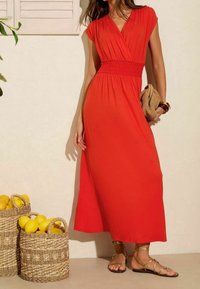 Abito maxi rosso con maniche corte e vita arricciata, realizzato in tessuto leggero. Abbinato a sandali dorati con cinturini e con una borsa intrecciata.