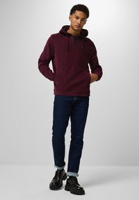 Burgundyröd hoodie med en framficka och dragkedja vid halsen, kombinerad med mörkblå jeans och svarta snörboots med kraftiga sulor.