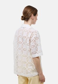 Chemise en dentelle blanche à manches courtes, avec un motif en découpes géométriques et un col à boutons. Tissu doux et léger.