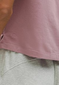 Camisa de manga corta en un tono mauve claro con un dobladillo redondeado y un logotipo en la parte inferior, superpuesta sobre pantalones cortos gris jaspeado con una costura visible.