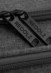 Graue Textiltasche mit zwei schwarzen Reißverschluss-Schlaufen, die den eingeprägten Text "REDOLZ" tragen. Nahaufnahme zeigt die gewebte Textur und die Details des Reißverschlusses.