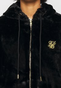 Sort hættetrøje med lynlås lavet af blødt, plush stof med et guld broderet logo og kontrasterende snore; har en hyggelig hætte og en glat lynlås.