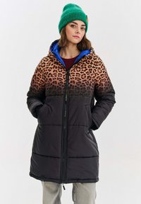 Žena v leopardom vzorovanom a čiernom prešívanom kabáte s kapucňou, so zelenou pletenou čiapkou a svetlošedými nohavicami, stojí so sladkami v vreckách.