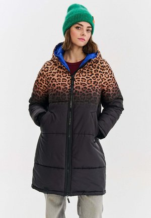 Žena v kabátu s leopardím potiskem a černé péřové bundě s kapucí, nosící zelenou pletenou čepici a světle šedé kalhoty, která stojí s rukama v kapsách.