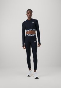 Črn športni crop top z zadrgo in dolgimi rokavi, kombiniran z legicami visoko pasu, ki imajo širok elastičen pas z blagovno znamko. Bele športne superge.