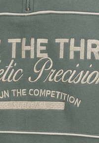 Grüner Pullover mit Reißverschluss am Kragen. Verziert mit cremefarbenen Stickereien: „BE THE THREE“ und „Athletic Precision“ sowie „SUBLEVEL.“