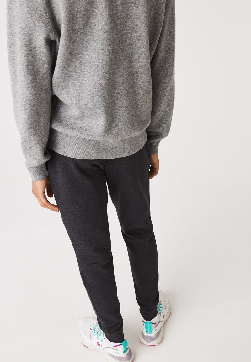 Persona che indossa un maglione grigio, pantaloni da jogging scuri e scarpe da ginnastica bianche con accenti turchesi e viola, in piedi davanti a uno sfondo bianco semplice.