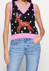 Schwarze Strickweste mit pinkem Kragen und Saum, verziert mit einem braunen Rentier und weißen Punkten. Über hellblauen Jeans getragen.