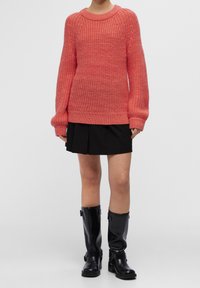 Osoba ubrana w grubą sweter koralowy, czarną plisowaną mini spódnicę i czarne wysokie buty za kolano zapinane na klamrę na jednolitym tle.