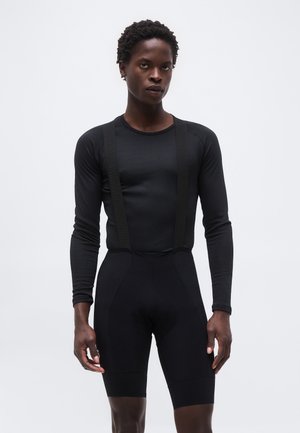 Mężczyzna ubrany w czarny, długi rękaw i czarne kolarskie spodenki z szelkami, stojący na tle jednolitego jasnoszarego tła.