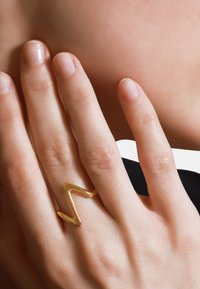 Goldener Zickzackring, der an einer Hand mit glatten, natürlichen Nägeln getragen wird. Der Ring zeichnet sich durch eine glänzende Oberfläche und ein offenes Design aus.