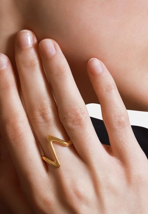 Goldener Zickzackring, der an einer Hand mit glatten, natürlichen Nägeln getragen wird. Der Ring zeichnet sich durch eine glänzende Oberfläche und ein offenes Design aus.