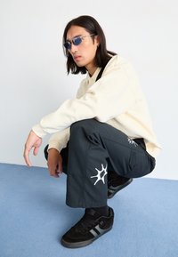 Pantaloni cargo negri cu desen alb pe fond, cămașă crem cu mânecă lungă și adidași negri. Ochelari de soare cu nuanțe albastre completează look-ul.