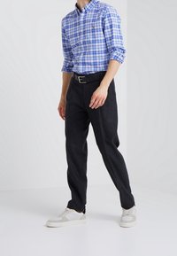 Blå och vit rutig skjorta med knappar, svart bälte, mörkgrå byxor och vita sneakers med grå detaljer, stående mot en ljus bakgrund.