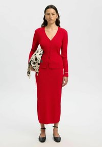 Cardigan rouge à côtes avec des manches longues et une fermeture à boutons, associé à une jupe à côtes assortie. Mis en valeur par une écharpe imprimée beige. Chaussures noires.