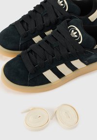 Tmavozelené semišové tenisky s čiernymi šnúrkami, obsahujúce tri béžové pruhy a logo na jazyku. Súčasťou sú aj dva okrúhle béžové šnúrky.