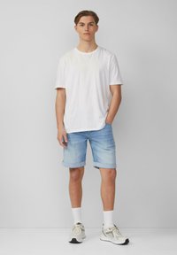 T-shirt en coton blanc, short en denim bleu clair avec ourlets retroussés, et baskets beiges avec accents gris. Fond uni, pose debout.