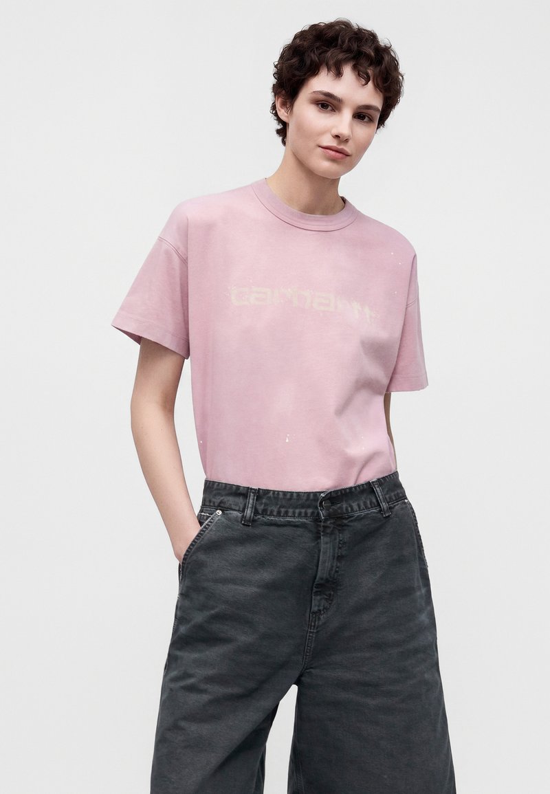 Junge Person mit kurzen lockigen Haaren, trägt ein ausgewaschenes pinkes T-Shirt und lockere dunkelgraue Jeans, steht mit den Händen in den Taschen.