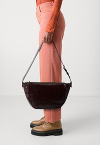 Borsa a tracolla in pelle bordeaux con una forma curvata, dotata di una tracolla nera e chiusura con zip, tenuta da una persona che indossa pantaloni e scarpe rosa.