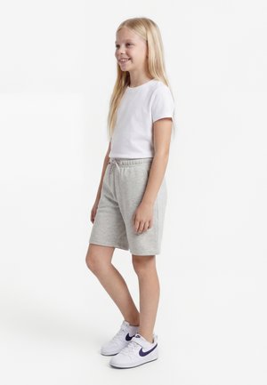 Κορίτσι με μακριά ξανθά μαλλιά στέκεται χαμογελαστή, φορώντας λευκό t-shirt, ανοιχτό γκρι σορτς και λευκά αθλητικά παπούτσια με μωβ σημάδια.