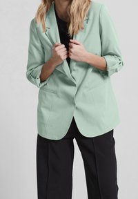 Mátově zelený blazer s jedním knoflíkem, klopovými límci a ohrnutými rukávy, zkombinovaný s černými kalhotami širokého střihu. Hladká textura.