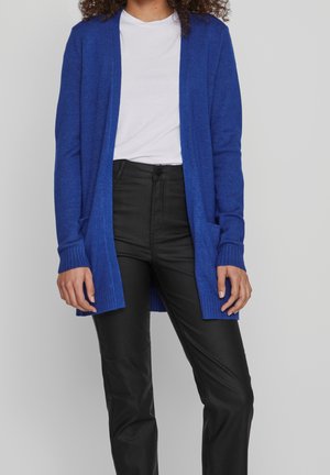 Femme portant un cardigan bleu à ouverture sur le devant, un t-shirt blanc et un pantalon noir taille haute, debout devant un fond clair uni.